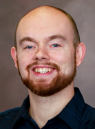 A man with a bald head, neatly trimmed beard, and mustache is smiling. He is wearing a dark shirt and looking directly at the camera against a neutral background.