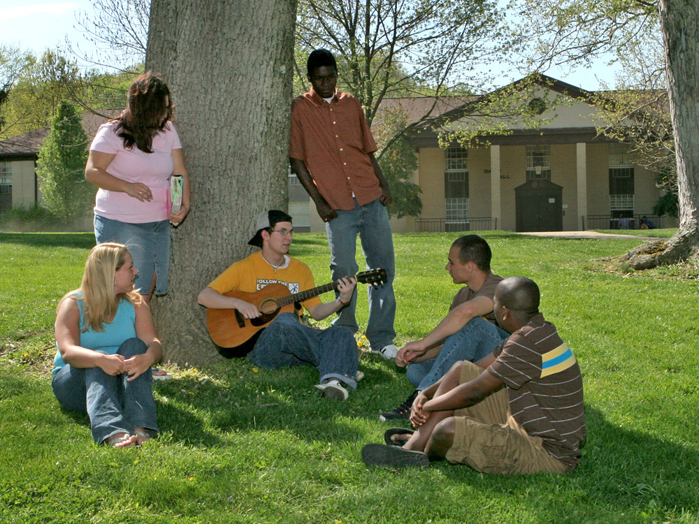 A group of six people sit and stand under a large tree. One person is playing a guitar, while others are listening and talking. A building is visible in the background.