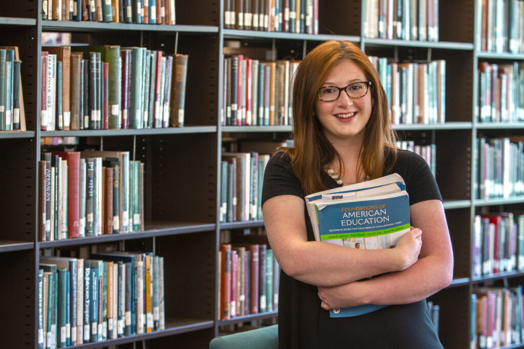 A woman with glasses stands in a library, smiling and holding a stack of books, including one titled "Foundations of American Education." She is dressed in casual attire.