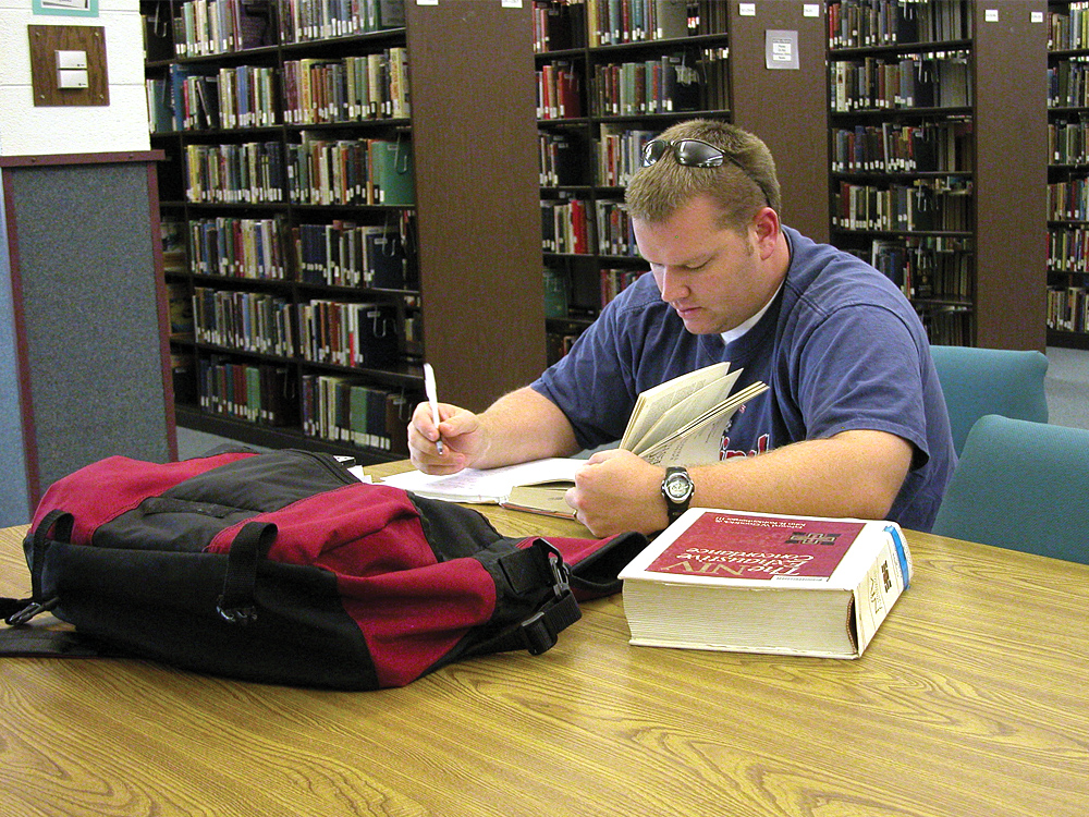 A person wearing a blue shirt is studying at a library table with a red and black backpack and an open book in front of them. Bookshelves are visible in the background.