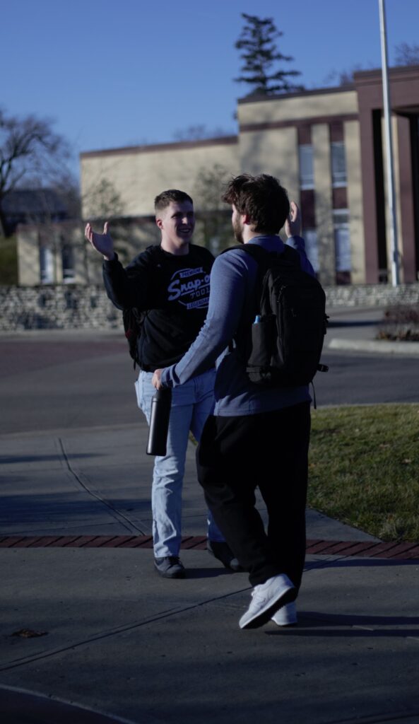 Two people with backpacks greet each other energetically on a sidewalk in front of a building. One person holds a bottle while gesturing with his arms.