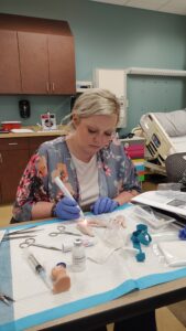 A person with gloves appears to be performing a medical procedure on a simulation model in a clinical setting, with various medical tools and equipment around them.