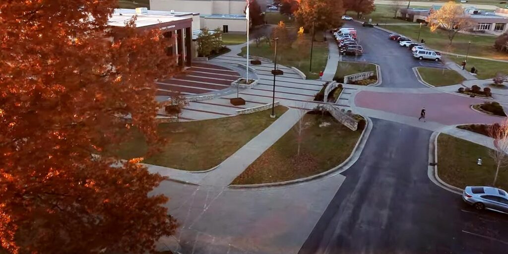 Aerial view of a modern building with a large parking lot, neatly landscaped areas, and scattered cars. The area is surrounded by autumn-colored trees.