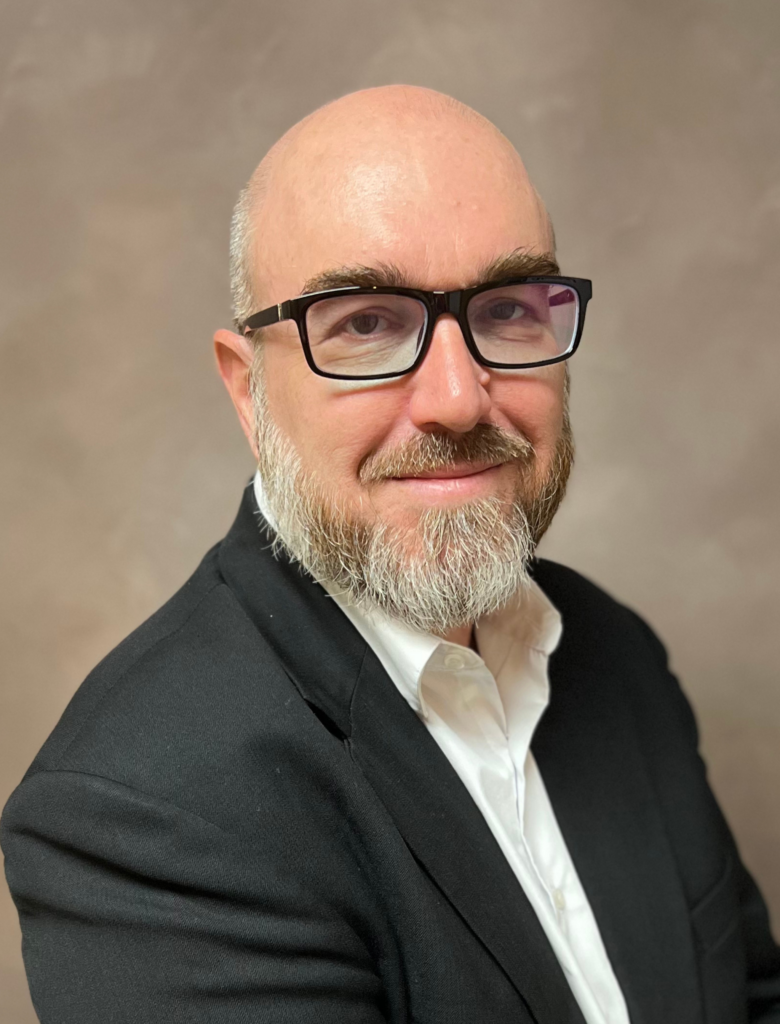 A bald man with glasses and a gray beard, wearing a black blazer and white shirt, poses against a plain background.