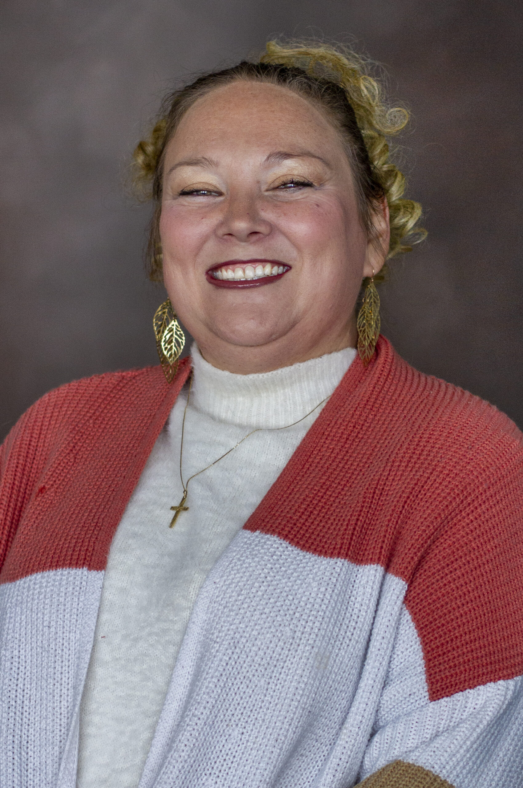 A person is smiling and wearing a red and white sweater, gold leaf earrings, and a gold cross necklace. The background is dark and neutral.