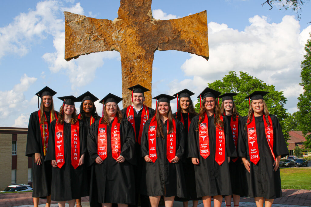 A group of nursing graduates in caps and gowns stands in front of a large cross outdoors on a sunny day. They wear red stoles with "Nursing" and "2024" written on them.