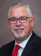 A man with short gray hair and glasses, wearing a black suit, white shirt, and red tie, smiles at the camera against a plain background.