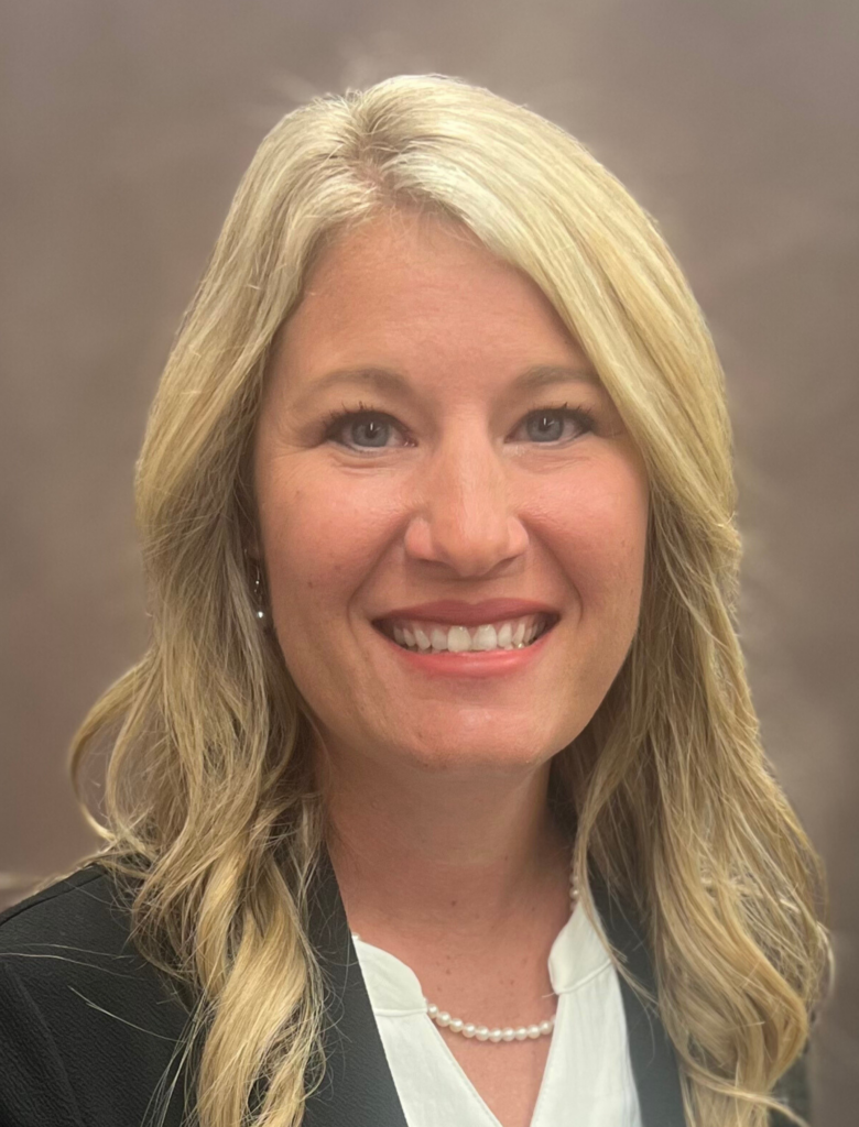 A woman with long blonde hair, wearing a black blazer, a white blouse, and a pearl necklace, smiles at the camera against a plain background.