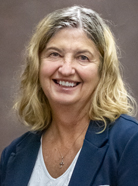 A woman with shoulder-length wavy blonde hair is smiling. She is wearing a dark blazer over a white top. The background is a plain brownish color.