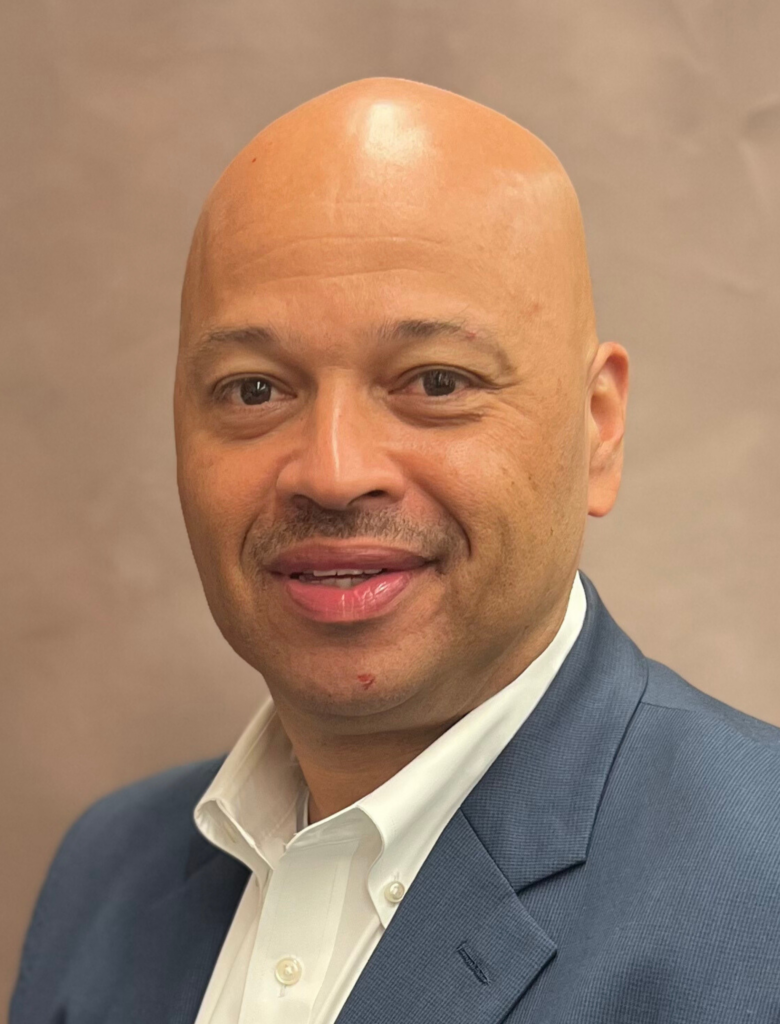 A bald man with a faint mustache and goatee wearing a navy blue blazer and white shirt smiles while looking at the camera. The background is a neutral brown color.