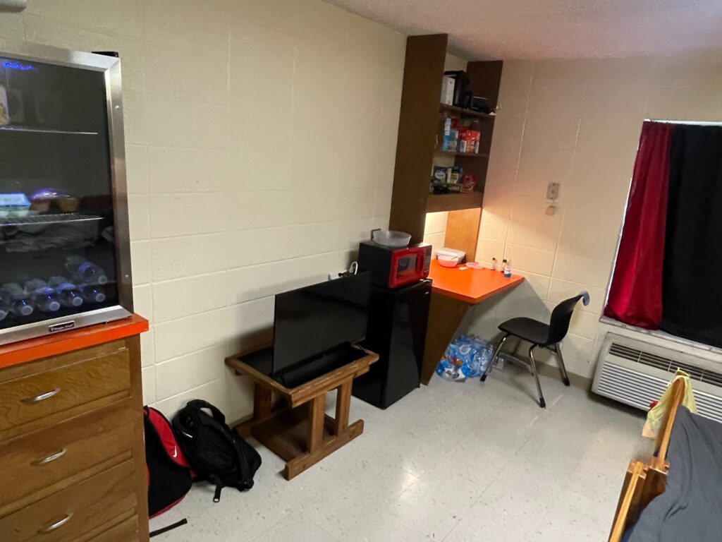 A small dorm room featuring a mini-fridge, microwave, desk with chair, TV on a stand, and various personal items. A backpack is on the floor, and a window with a curtain is partially visible.