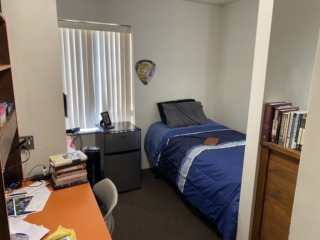 A small bedroom with a single bed, a mini fridge, a desk with books and papers, and a shelf of books. Vertical blinds cover the window.