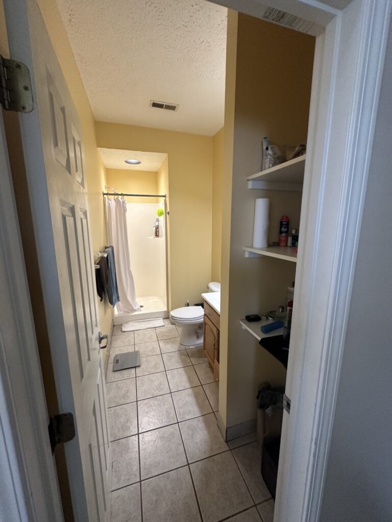 A small bathroom with a shower, toilet, and sink. Open shelves on the right contain various items. The walls are yellow, and the floor has white tiles. A white door is partially open.