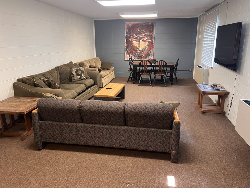 A living room with two sofas, a coffee table, side tables, a wall-mounted TV, and a dining table with six chairs. A colorful abstract painting hangs on the wall behind the dining table.