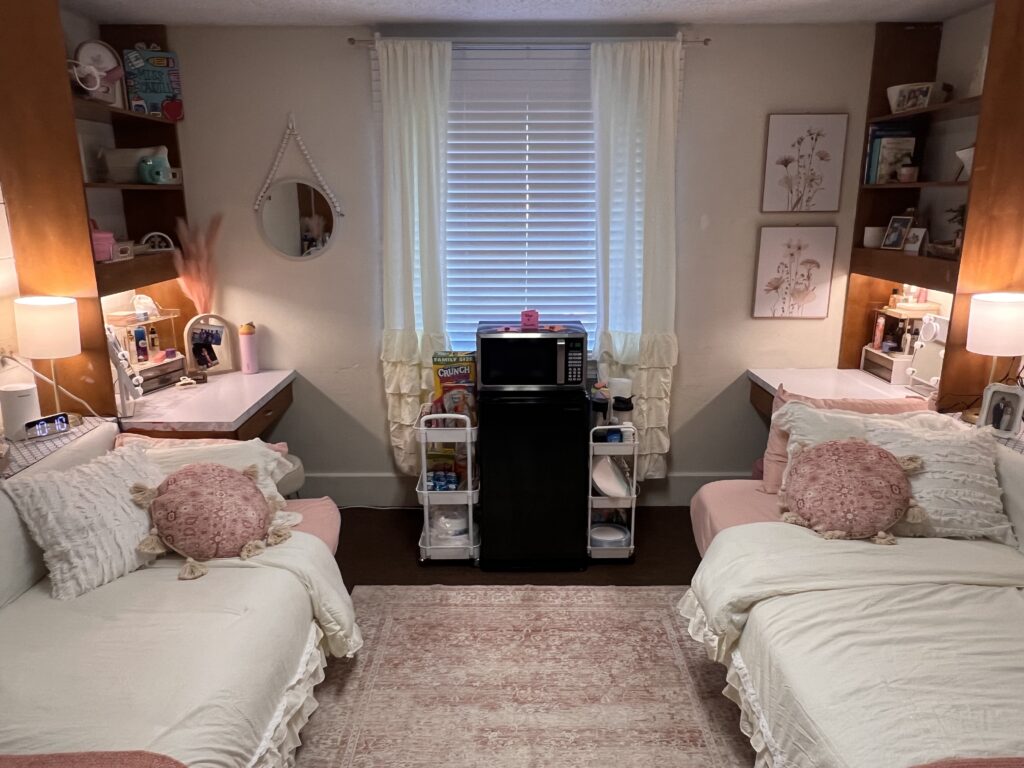 A symmetrical dorm room with two twin beds, each accompanied by a desk and shelves. A window with a blind is centered between the beds, and a small refrigerator and microwave stand below the window.