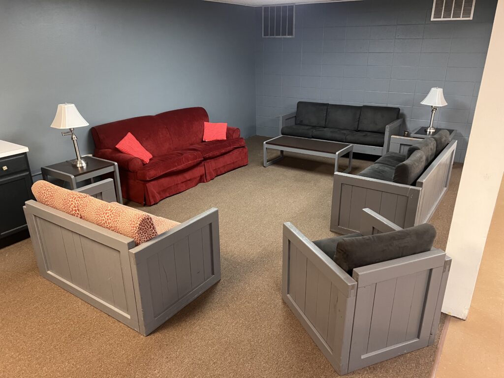 A room with a brown carpet, gray walls, and six upholstered seats arranged in a square formation around a central space, two lamps on tables beside the red sofa, and minimal additional furnishings.