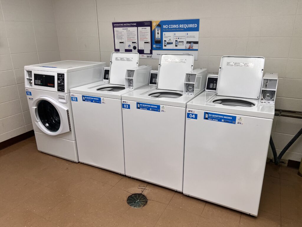 A laundromat with three top-loading washing machines and one front-loading washing machine, with instructions on a wall sign above, indicating that no coins are required.