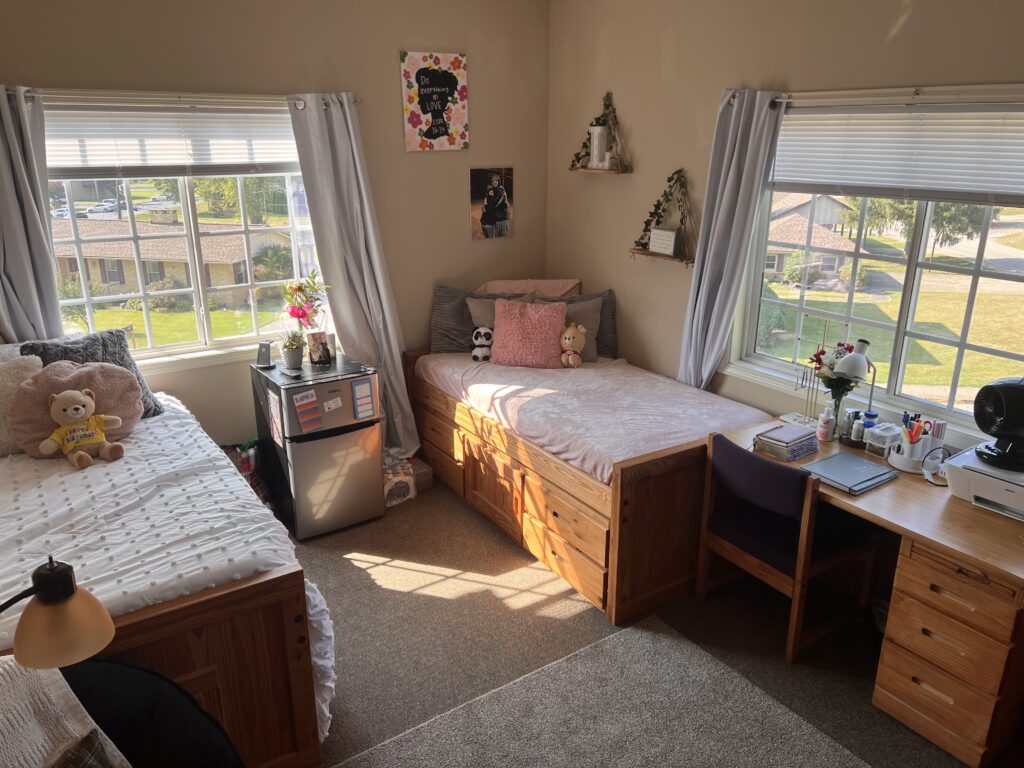 A tidy bedroom with two single beds, two desks, decorations on the walls, and natural light from two windows illuminating the room.