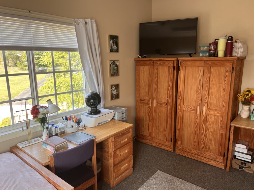 A well-organized room with a wooden desk and chair, a wooden wardrobe, a TV mounted on top of the wardrobe, and a window with white blinds. Various items are neatly arranged.