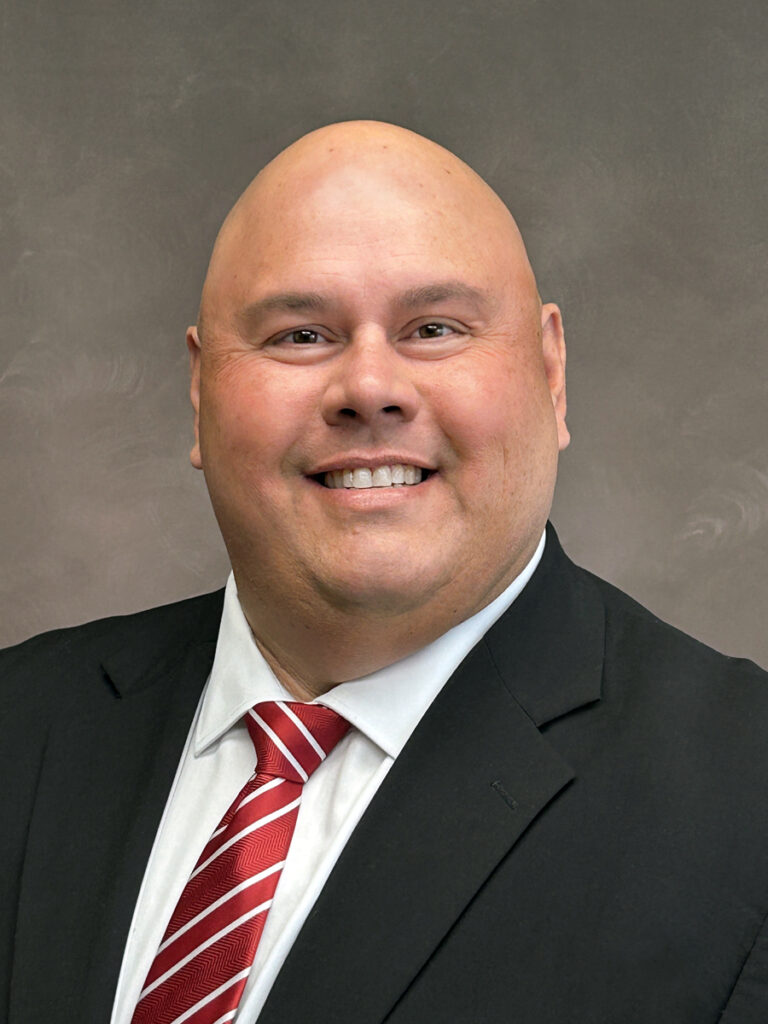 A bald man in a black suit jacket, white shirt, and red striped tie smiles at the camera against a neutral background.