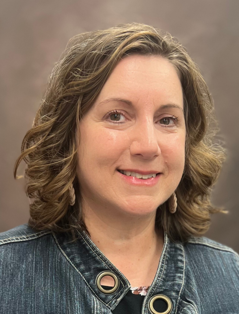 A woman with short curly hair, wearing a denim jacket and earrings, smiles at the camera against a plain background.