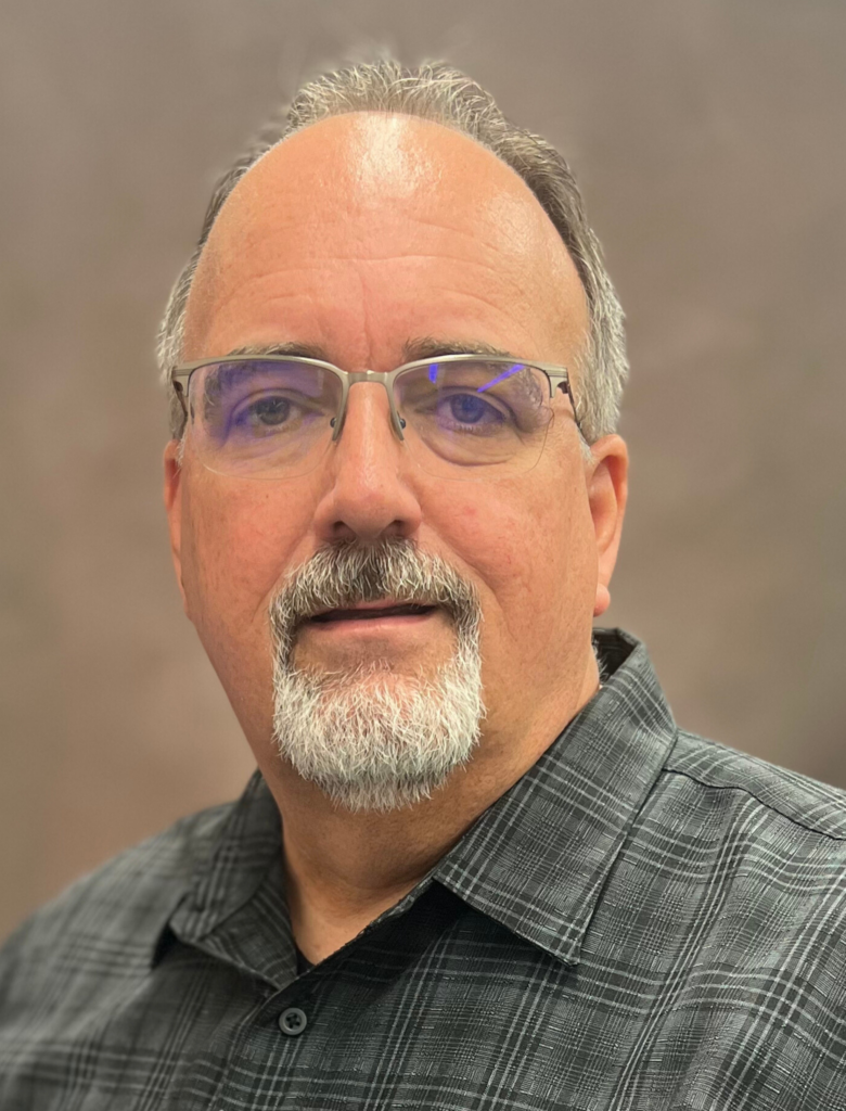 A man with short gray hair and a beard, wearing glasses and a gray plaid shirt, poses against a neutral background.