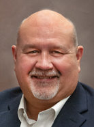 A balding man with a gray beard, wearing a dark suit jacket over a white shirt, smiles at the camera against a plain, muted background.