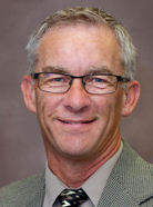 A man with short gray hair, glasses, and a greenish-gray blazer smiles at the camera against a plain background.