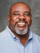 A man with a beard and a smile is wearing a light blue collared shirt against a neutral background.