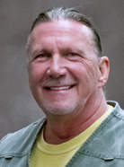 A man with slicked-back hair and a visible smile, wearing a green collared shirt over a yellow top, is posing in front of a grey background.