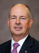 Middle-aged man with short hair in a suit and red tie, standing against a plain brown background.