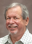 A man with gray hair and a beard is smiling at the camera. He is wearing a light-colored, checkered shirt and standing against a plain, neutral background.