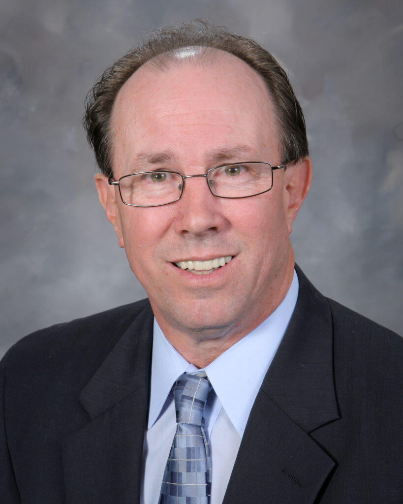 A middle-aged man with glasses, wearing a suit and tie, poses for a professional portrait against a gray backdrop.