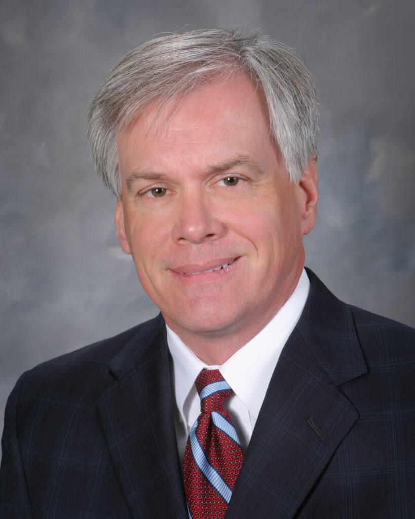 A man with gray hair wearing a dark suit, white shirt, and red-striped tie, poses against a neutral backdrop.