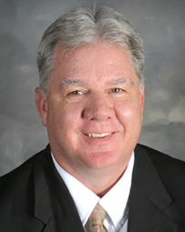 A man with short gray hair, wearing a dark suit and a light-colored tie, smiles at the camera against a neutral background.