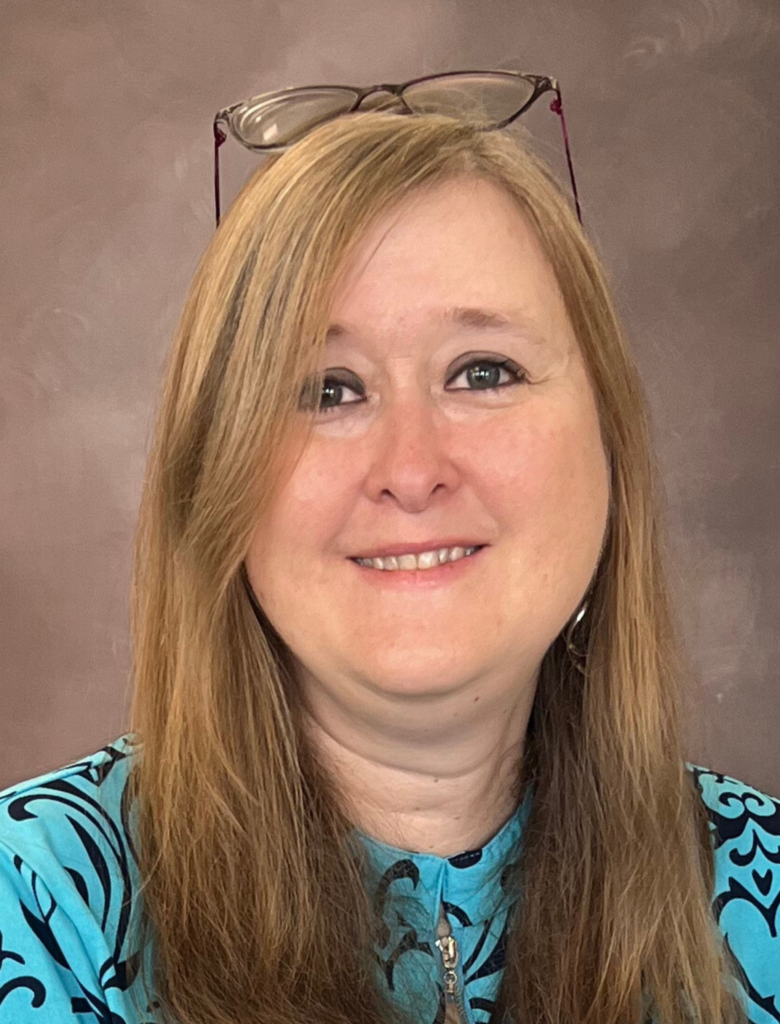 A woman with long, light brown hair smiles at the camera. She is wearing glasses on her head, a turquoise patterned top, and has a neutral brown background behind her.