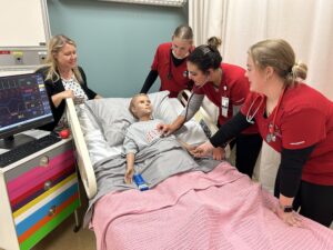 Four nursing students in red uniforms practice on a medical mannequin in a hospital bed, with a heart rate monitor nearby. An instructor observes the session.