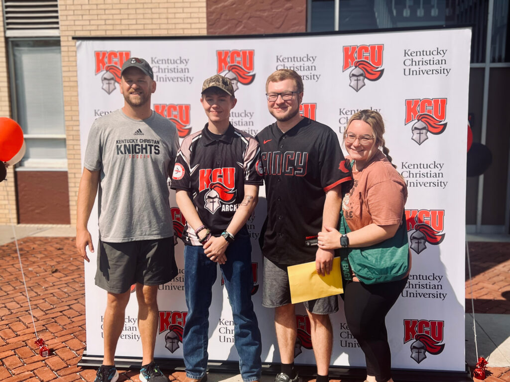 Four people stand smiling in front of a Kentucky Christian University backdrop outdoors, wearing university-themed shirts and casual clothing.