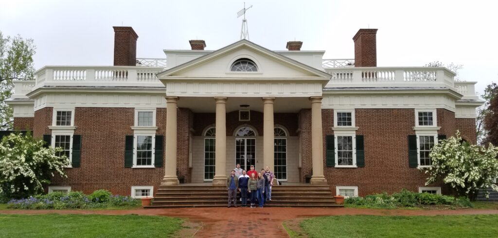 A group of people stand in front of a historic two-story brick mansion with white columns and green shutters under a cloudy sky.