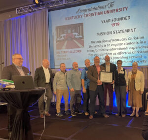 A group of people on stage hold a framed certificate with a congratulatory message to Kentucky Christian University displayed on a screen in the background.